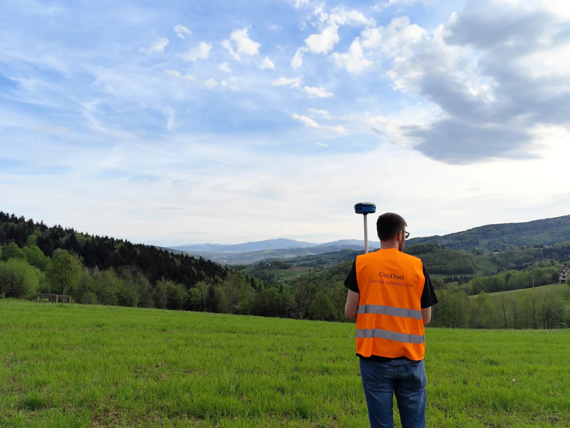 Geodeta GeoDuet wykonujący pomiar odbiornikiem GNSS na tle pagórkowatego terenu